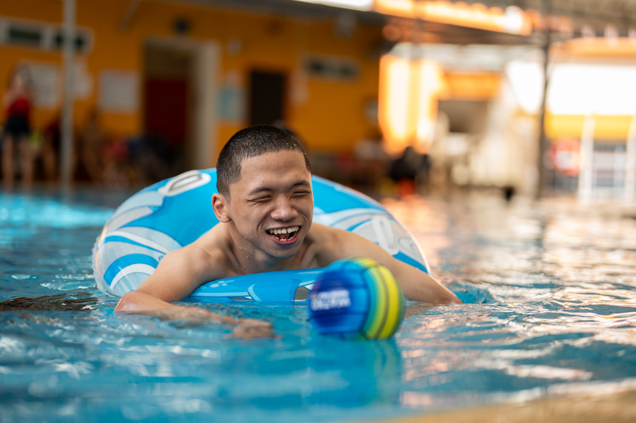 Person in pool, smiling, floating in blue inflatable ring with a ball.